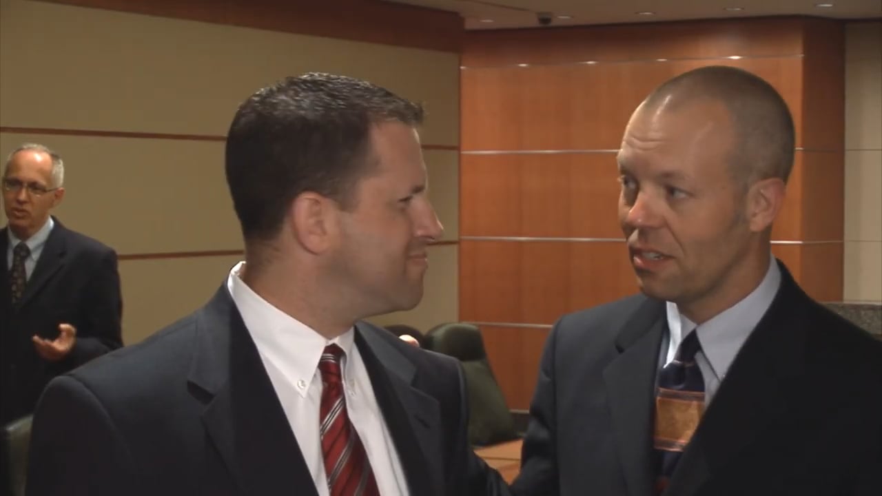 3 men in suits are standing around a Board Room table. In the foreground, one man talks to another who reacts with confusion.